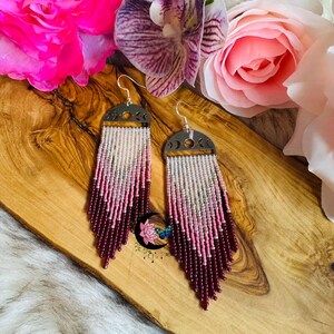 May include: A pair of beaded earrings with a silver-tone crescent moon design. The earrings feature a gradient of white, pink, and burgundy beads, creating a fringe effect. The earrings are displayed on a wooden surface with flowers.