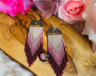 May include: A pair of beaded earrings with a silver-tone crescent moon design. The earrings feature a gradient of white, pink, and burgundy beads, creating a fringe effect. The earrings are displayed on a wooden surface with flowers.