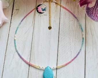 May include: A beaded choker necklace with a gradient of colors, including pink, purple, blue, and yellow. The necklace features a teardrop-shaped turquoise pendant and a gold-colored chain.