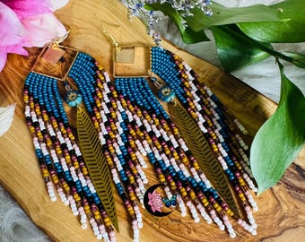 May include: A pair of handcrafted earrings with a bohemian style. The earrings feature a square gold-colored top, blue beaded section, and long fringe in white, yellow, and burgundy. A gold-colored leaf charm is attached to the fringe. The earrings are displayed on a wooden surface.