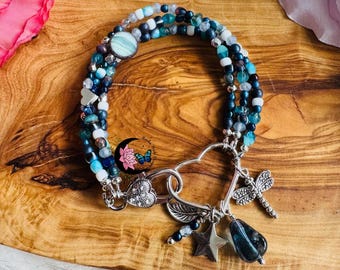 May include: A multi-strand bracelet with blue, white, and silver beads. The bracelet features a heart-shaped clasp and charms including a dragonfly, star, and teardrop-shaped stone. The bracelet is displayed on a wooden surface.