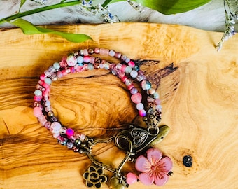 May include: A multi-strand beaded bracelet in shades of pink, purple, and gold. The bracelet features a turtle clasp, a pink flower charm, and a gold-colored heart and flower charm. The beads are various sizes and shapes.