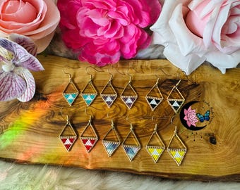 May include: A collection of handmade beaded earrings with gold-colored frames. The earrings feature geometric diamond shapes with various color patterns, including turquoise, white, black, red, and yellow. The earrings are displayed on a wooden surface with flowers.