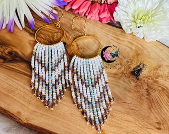 May include: A pair of handmade beaded earrings. The earrings feature a gold-colored hoop with a clover-shaped top. The beaded fringe is white, light blue, and purple, with gold-colored beads at the bottom. The earrings are displayed on a wooden surface.