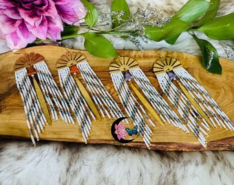 May include: Four pairs of handmade beaded earrings. Each pair features a wooden sunburst design, white and blue beads, and a gold-colored metal accent. The earrings are displayed on a wooden surface with floral and leaf accents.