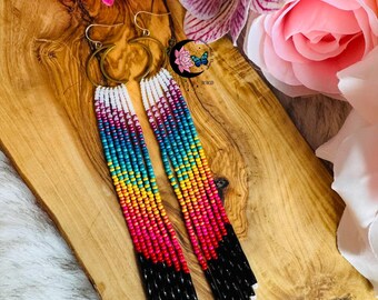 May include: Handmade beaded earrings with a vibrant rainbow gradient design. The earrings feature gold-colored hoops and long, beaded fringe in shades of white, blue, yellow, pink, red, and black. The earrings are a unique accessory.