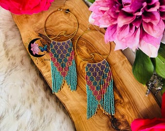 May include: Gold hoop earrings with turquoise, purple, pink, and gold beaded fringe. The earrings are displayed on a wooden board with pink and red flowers in the background. The earrings have a wave design.