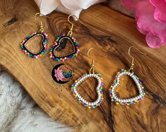 May include: Gold-tone heart-shaped earrings with colorful beaded edges. The earrings have a hollow center and are displayed on a wooden surface. The earrings are a unique accessory.