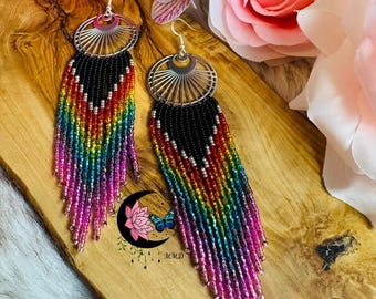 May include: A pair of handmade earrings with a silver fan-shaped top and long beaded fringe in a rainbow of colors. The top section is black, transitioning to red, orange, yellow, green, blue, and purple. The earrings are displayed on a wooden surface.