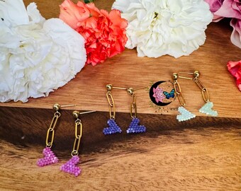 May include: Four pairs of gold heart-shaped earrings with beaded details. The earrings are displayed on a wooden surface with flowers in the background.