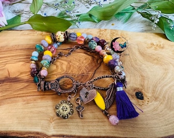 May include: A multi-colored beaded bracelet with a copper-toned clasp and charms. The bracelet features a variety of beads in shades of yellow, pink, green, and purple. Charms include a heart, key, and tassel. The background is a wooden surface.