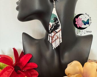 May include: Handmade beaded earrings with a black, white, teal, and brown design. The earrings are displayed on a black mannequin head. The earrings have a fringe at the bottom. The earrings are a unique accessory.