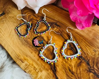 May include: Heart-shaped earrings with beaded edges in various colors. The earrings have a silver hook and are displayed on a wooden surface. The earrings are a fashion accessory.