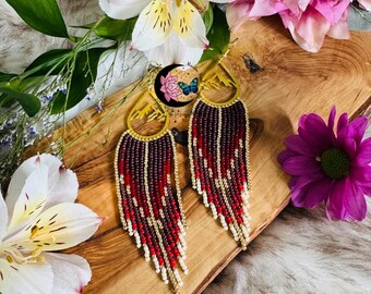 May include: A pair of handmade beaded earrings with a gold-colored hoop and long fringe. The fringe features a pattern of red, white, and brown beads. The earrings are displayed on a wooden surface with flowers.