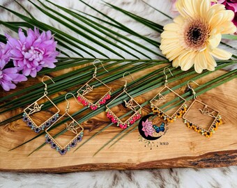 May include: Four pairs of gold-toned earrings with square frames and dangling beads. The earrings feature a small origami bird charm. The beads are in shades of purple, pink, red, and yellow.