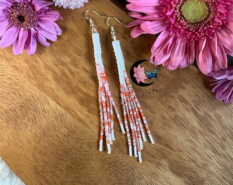 May include: A pair of long, beaded earrings with white and orange beads. The earrings are hanging from silver hooks.