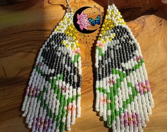 May include: Handmade beaded earrings with a unique design. The earrings feature a combination of white, black, green, pink, and yellow beads. The design includes a floral pattern and a fringe at the bottom. The earrings are displayed on a wooden surface.
