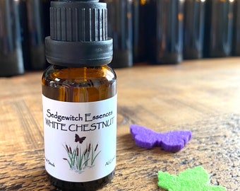 May include: A brown glass bottle with a black dropper, labeled "Sedgewitch Essences WHITE CHESTNUT." The label features a butterfly and cattails illustration. A purple butterfly, green leaf, and decorative key are nearby.