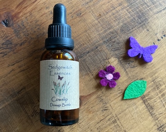 May include: A brown glass bottle with a black dropper, labeled "Sedgewitch Essences Cowslip Dosage Bottle." Beside it are felt decorations: a purple butterfly, a purple flower, and a green leaf. The items are arranged on a wooden surface.