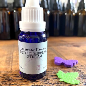 Op de afbeelding: Een kobaltblauw glazen druppelflesje met een witte dop en een etiket met de tekst Sedgewitch Essences BE THE BURBLING STREAM en een gulden snede spiraalafbeelding. Ernaast liggen een paarse vilten vlinder, een groen vilten blaadje en een bronzen sleutel.
