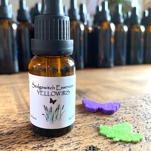 May include: A brown glass bottle with a black dropper, labeled "Sedgewitch Essences YELLOW IRIS." The label features a butterfly and cattails illustration. A purple butterfly and green leaf-shaped felt cutouts and a decorative key are nearby.