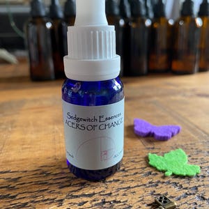 Op de afbeelding: Een kobaltblauw glazen druppelflesje met een witte dop en een wit etiket met de tekst Sedgewitch Essences ACERS OF CHANGE en een gulden snede spiraalafbeelding. Het flesje staat bij een paarse vilten vlinder en een groen vilten blaadje.