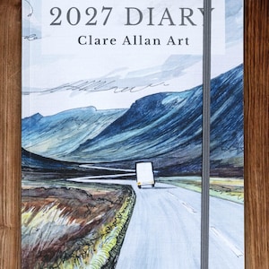 May include: A 2027 diary by Clare Allan Art featuring a landscape illustration of a road winding through blue mountains with a white vehicle driving forward. The cover has a light blue sky, dark blue hills, and a vertical grey elastic closure band.