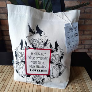 Może przedstawiać: Kremowa torba typu tote z czarnymi uchwytami i grafiką postaci z kreskówki. Tekst na torbie brzmi: "I'M YOUR GUY, YOUR DAY-TO-DAY, YOUR CHUM, YOUR STEADFAST HOTELIER." Dołączona jest metka ze słowem "Neutral".