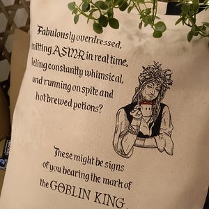 May include: Cream tote bag with black text and a black illustration of a crowned figure. The text reads "Fabulously overdressed, emitting ASMR in real time...the GOBLIN KING."