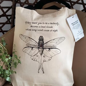 Puede incluir: Bolso tote de color crema con una ilustración de polilla en blanco y negro. Contiene la cita: "If they want you to be a butterfly, become a loud cicada whose song won't cease at night." Una etiqueta dice "We Believe in Being Okay."