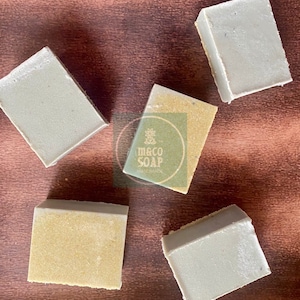 May include: Five handmade soap bars in shades of green and yellow are arranged on a brown surface. The soaps are rectangular with a textured appearance. The image includes a logo that reads "m&co SOAP HANDMADE".
