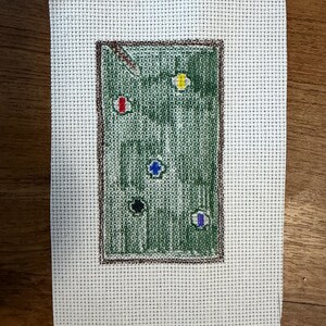 May include: A rectangular piece of white Aida cloth featuring a needlepoint design of a green pool table with a brown border. The design includes a pool cue and several colored balls in red, yellow, blue, black, and white scattered on the surface.