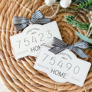 May include: Two white Christmas ornaments with black and white gingham ribbon. The ornaments feature the text "Christmas - 2023" and a number, "75423" and "75490". The ornaments also include the names "Celeste" and "Trenton" and the words "Home" and "Texas".