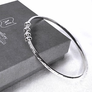 May include: A silver bangle bracelet with a hammered finish and four small silver beads. The bracelet is displayed on a gray box with the text "AAM Jewellery" printed on it.