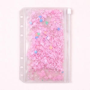 May include: A clear, rectangular, zippered pouch filled with pink confetti, including shell and butterfly shapes. The pouch has a row of holes along the left side for binding. The confetti is a mix of pink hues and small colorful accents.