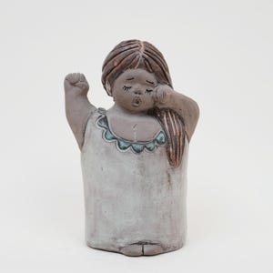 Puede incluir: Figura de cerámica de una niña con pelo largo castaño, bostezando y frotándose el ojo. Lleva un vestido gris claro con ribete turquesa. La escultura tiene un aspecto envejecido y desgastado.