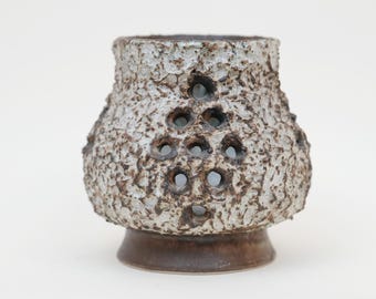 Könnte beinhalten: Eine strukturierte Keramikvase mit hellbraunem Sockel und einer rauen, gesprenkelten Außenseite in Grau- und Brauntönen. Die Vase hat mehrere kreisförmige Öffnungen, die ein einzigartiges Designelement hinzufügen.