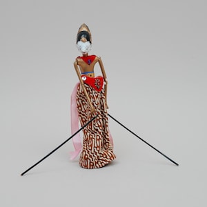 Könnte beinhalten: Eine traditionelle indonesische Wayang Golek-Puppe. Die Puppe hat ein bemaltes Gesicht, eine Kopfbedeckung und ist in einem rot-braunen gemusterten Kostüm gekleidet. Zwei schwarze Stäbe sind an der Puppe befestigt.