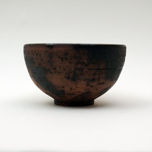 May include: A small, dark brown ceramic bowl with a rough, textured surface. The bowl has a slightly rounded shape and a wide, open rim.