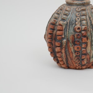 Puede incluir: Un pequeño jarrón redondo de cerámica con una superficie texturizada. El jarrón está decorado con patrones marrones, verdes y blancos.