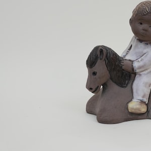 Pode incluir: Uma figurinha de cerâmica de uma criança a andar a cavalo. A criança está vestida de branco e tem pele morena. O cavalo é castanho e tem uma crina preta.