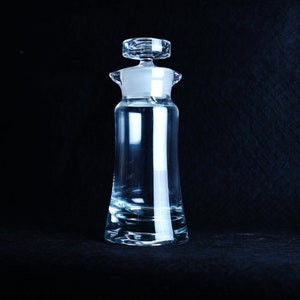 Puede incluir: Una botella de vidrio transparente con un tapón de vidrio. La botella tiene una base redondeada y un cuello estrecho. El tapón tiene una parte superior plana y una pequeña perilla.