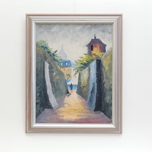 May include: A framed oil painting depicting a narrow street scene with buildings on either side. The street is lined with trees and a single figure walks down the center. The painting is done in a muted palette of blues, greens, and yellows.