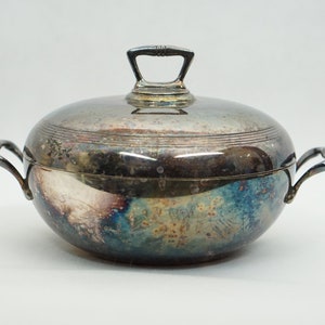 Puede incluir: Una fuente de servir de plata con tapa y dos asas. La fuente es redonda y tiene un acabado brillante. La tapa tiene una pequeña asa en la parte superior.