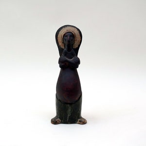 May include: A black ceramic figurine of a person with a white halo, standing with arms crossed, wearing a dark robe and green pants.
