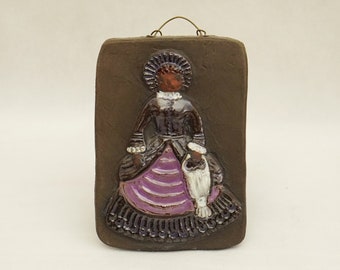 May include: A ceramic wall plaque featuring a woman in a purple and brown dress with a white hat. The plaque has a brown background and a metal hanger on the top.