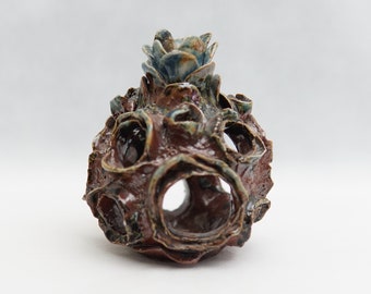 Könnte beinhalten: Eine braune Keramikskulptur mit einer strukturierten Oberfläche und einer blauen und grünen Blume oben. Die Skulptur hat viele Löcher und scheint hohl zu sein.