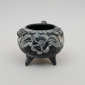 May include: A small, round, black ceramic vessel with three short legs and a wide opening. The exterior surface features white etched floral patterns and two small sculpted animal-like figures positioned on the upper rim of the bowl.