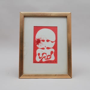 Könnte beinhalten: Ein gerahmtes Kunstwerk mit einem rot-weißen abstrakten Druck, der eine kreisförmige obere Form und eine fließende, organische untere Form zeigt. Der Druck enthält einen roten Spritzereffekt und handschriftlichen Text. Der Rahmen ist goldfarben.