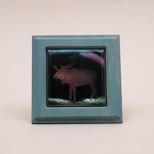 May include: A square decorative tile framed in blue-grey wood. The tile features a dark, glossy surface with a silhouette of a moose in a reddish-brown hue. Above the moose is a purple streak, and below it is a patch of light green texture.
